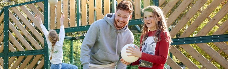 Decoratieve foto van een coach Gezonde Kinderopvang. Naast hem staan een meisje dat een bal vasthoudt. De coach en het meisje kijken naar hetzelfde en lachen samen.