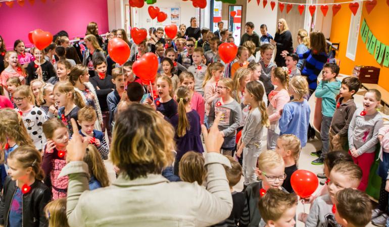 Decoratieve foto van basisschoolleerlingen. Samen met hun leraren zingen ze een lied. Ze houden ballonnen in de vorm van een hart vast.